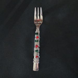 Puede incluir: Un tenedor de metal plateado con el mango envuelto en alambre de plata decorativo. El alambre enrollado incorpora pequeñas cuentas rojas y verdes alternadas a lo largo del mango, creando una apariencia texturizada y colorida en el utensilio.