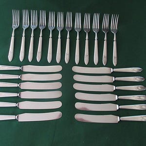 May include: A set of twelve silver-toned metal forks and twelve silver-toned metal knives arranged in two rows. Each fork features four tines and a slender handle. Each knife has a smooth, rounded blade and a handle with a decorative pattern.