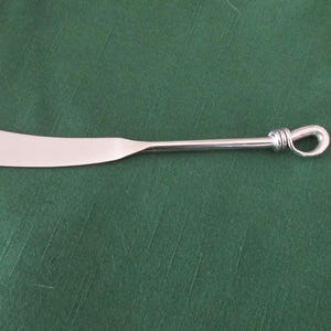 May include: A stainless steel butter knife with a curved blade and a looped handle. The knife is shown on a green background. The handle has a decorative knot design.