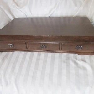 May include: A rectangular, dark brown wooden box with three small drawers. The drawers have ornate, dark metal handles. The box is set on a white and gray striped fabric.