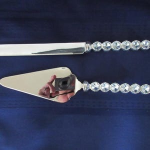 May include: A silver cake knife and server set. The knife and server have a shiny, reflective surface. The handles are decorated with a row of clear, faceted crystal-like stones. The set is displayed against a dark blue background.