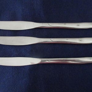 May include: Three silver-toned metal dinner knives arranged horizontally. Each knife features a smooth, rounded blade and a handle decorated with a subtle, engraved curved line and two small, four-pointed starburst patterns near the end.