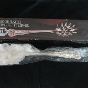 May include: A silver-plated spaghetti server in its original box. The box is dark with the words "Silver Plated Spaghetti Server" in white. The server is in a plastic bag. The server has a decorative handle and a slotted head.