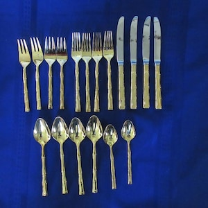 Könnte beinhalten: Ein goldfarbenes Besteckset auf einer blauen Oberfläche. Das Set umfasst Gabeln, Messer und Löffel. Die Griffe haben ein bambusartiges Design. Die Anordnung deutet auf eine formelle Essumgebung hin.