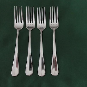 May include: A set of four polished silver-toned metal forks arranged in a vertical row. Each fork features a long, slender handle that widens slightly at the base and four tines. The metal surface is smooth and reflective, showing a metallic sheen.