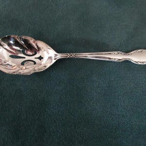 Può includere: Un cucchiaio da portata d'argento con manico decorativo e coppetta smerlata e fessurata. Il cucchiaio presenta un design ornato sul manico e sulla coppetta, su sfondo verde scuro. Il cucchiaio è probabilmente in argento sterling.