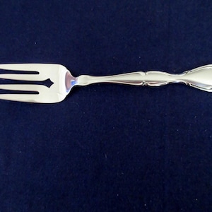 Può includere: Una forchetta d'argento con tre rebbi e un manico decorativo. La forchetta ha un ritaglio a forma di diamante tra i rebbi. Il manico presenta un design dettagliato e ornato. La forchetta è posta su uno sfondo blu scuro.