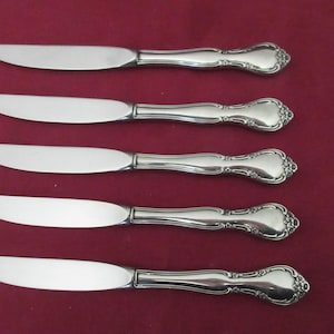 May include: Five silver-colored butter knives with ornate handles are arranged on a burgundy background. The blades are smooth and curved, while the handles feature a decorative floral design. The knives are likely part of a silverware set.