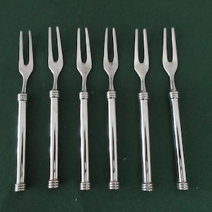 May include: Six silver serving forks with three tines each, arranged in a row. The forks have long, slender handles with a decorative, ridged design near the base. The background is a solid, dark green.