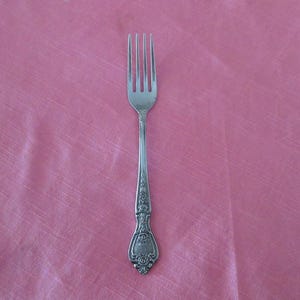 May include: A silver fork with four tines and a decorative handle. The handle features floral and scrollwork designs. The fork is set against a pink background.