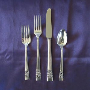 Puede incluir: Un juego de cuatro utensilios plateados: un tenedor, un tenedor de ensalada, un cuchillo y una cuchara. Los utensilios están dispuestos verticalmente sobre un fondo morado. Cada pieza tiene un diseño floral decorativo en la base del mango.