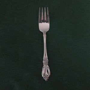 May include: A silver fork with four tines and an ornate handle. The fork is centered on a dark green background. The handle features a detailed floral design.