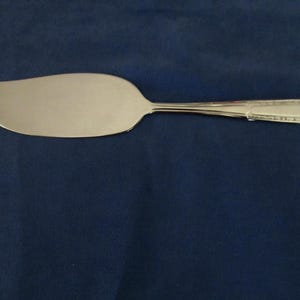 May include: A silver-colored pie server with a wide, curved blade and a decorative handle. The server is made of a shiny metal and is set against a dark blue background. This kitchen utensil is designed for serving pies and cakes.