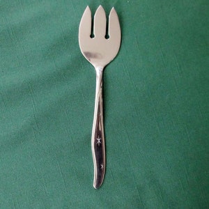 May include: A silver serving fork with three tines and a decorative handle. The handle has starburst designs. The fork is set against a green background.