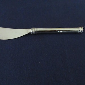May include: A silver butter knife with a rounded blade and a cylindrical handle. The handle has a ribbed design near the blade and at the end. The knife is set against a dark blue background.