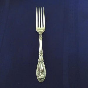 May include: Silverware fork with a decorative handle featuring a grape cluster design. The fork has four tines and is set against a dark blue background. The fork is made of a shiny, reflective metal.