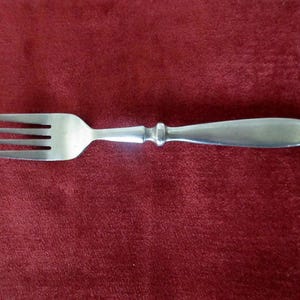 May include: A silver-colored metal fork featuring four tines, a smooth neck with a decorative rounded collar, and a solid, elongated handle with a rounded end. The utensil has a polished finish and a simple, clean design throughout its structure.