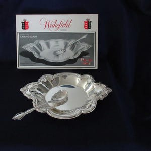 May include: A silver-plated bowl and spoon set featuring the Wakefield pattern. The packaging displays the text Wakefield Pattern, International DeepSilver, and Two Piece Party Set. The bowl has a scalloped edge and a polished reflective finish.