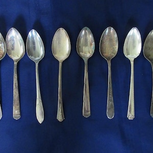May include: A collection of eight silver-toned metal spoons arranged in a horizontal row. Each spoon features a unique handle design with varying degrees of tarnish and patina, showcasing different decorative patterns and shapes on the stems.