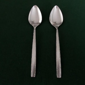 May include: Two silver spoons with oval bowls and detailed handles are displayed against a dark green background. The spoons are positioned vertically, showcasing their polished surfaces and intricate design. The spoons are likely for serving or eating.