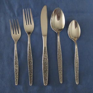Puede incluir: Un juego de cinco cubiertos plateados: dos tenedores, un cuchillo y dos cucharas. Cada mango está decorado con un diseño floral. Los cubiertos están colocados sobre una superficie azul.