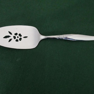 May include: A silver pie server with a floral cutout design on the blade. The handle has a decorative pattern near the top. The server is set against a dark green background.