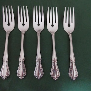 Può includere: Cinque forchette d'argento con manici decorati e un intaglio a forma di diamante nei rebbi. Le forchette sono disposte su una superficie verde scuro, evidenziando il loro design elegante. Ideali per cene formali.