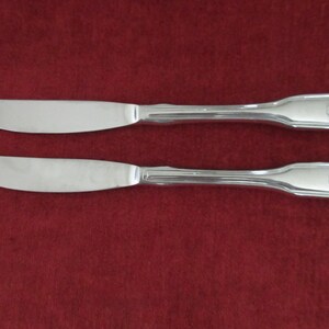 May include: Two silver-toned metal dinner knives with smooth, rounded blades and handles featuring a vertical grooved pattern and a decorative shell motif at the base. The knives have a polished, reflective finish and a classic, symmetrical design.