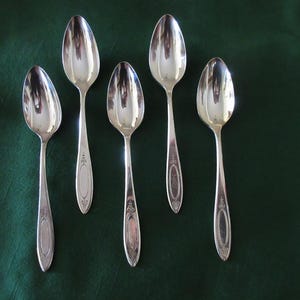 May include: Five silver spoons arranged on a dark green surface. The spoons have a classic design with elongated handles and decorative detailing. The spoons are reflective, showing the surrounding environment.
