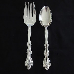 Può includere: Una forchetta e un cucchiaio d'argento con manici decorati. La forchetta ha quattro denti e il cucchiaio ha una ciotola arrotondata.