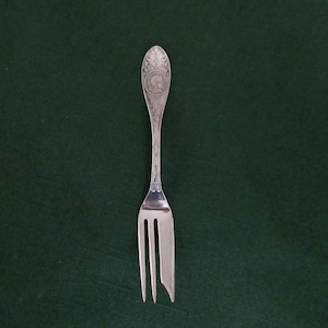 Puede incluir: Un tenedor plateado con tres púas y un mango ornamentado. El mango presenta un diseño detallado, posiblemente un escudo. El tenedor se encuentra sobre un fondo verde oscuro, resaltando su superficie pulida. Una pieza clásica de cubertería.