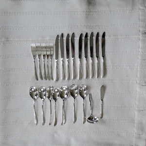 May include: A set of silverware, including forks, knives, and spoons, laid out on a white tablecloth. The silverware is made of polished silver and has a traditional design.