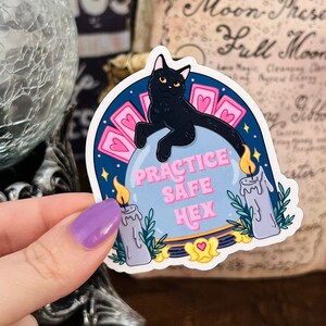 Puede incluir: Una pegatina con un gato negro sentado en una bola de cristal con el texto "Practice Safe Hex" en letras rosas. La bola de cristal está rodeada de corazones rosas y dos velas moradas.