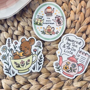May include: Three stickers with tea-themed designs are displayed on a woven mat. One sticker features a mouse reading in a teacup. Another says "Tea fixes everything." The third says "Powered by Tea & To-Do Lists."