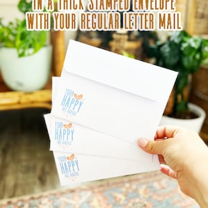 K&ouml;nnte beinhalten: Drei wei&szlig;e Umschl&auml;ge mit einem blauen und orangefarbenen Logo, auf dem "Your Happy Has Arrived" steht. Die Umschl&auml;ge sind &uuml;bereinander gestapelt und werden von einer Hand gehalten. Der Text "Stickers are delivered in a thick stamped envelope with your regular letter mail" ist im Hintergrund sichtbar.