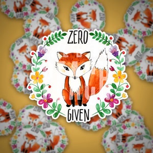 Pode incluir: Um adesivo branco com uma ilustra&ccedil;&atilde;o em aquarela de uma raposa com uma coroa de flores ao redor. O texto "Zero F***s Given" est&aacute; escrito no adesivo.