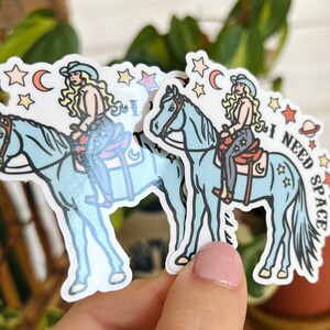 May include: Two blue horse stickers with a woman riding each horse. The woman is wearing a cowboy hat and a blue and white outfit. The sticker on the right has the text "I NEED SPACE" on it.