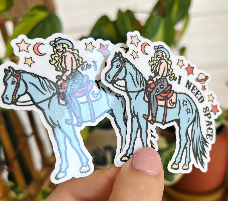 May include: Two blue horse stickers with a woman riding each horse. The woman is wearing a cowboy hat and a blue and white outfit. The sticker on the right has the text "I NEED SPACE" on it.