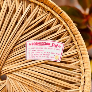 Puede incluir: Pegatina rosa y blanca con el texto "*PERMISSION SLIP*" y una lista de declaraciones de permiso: "To feel however you want to feel", "To make mistakes", "To not figure it all out right away", "To be kinder to yourself", "To take all the time you need". Una carita sonriente est&aacute; al final de la &uacute;ltima declaraci&oacute;n.