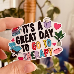 May include: A white sticker with the text "IT'S A GREAT DAY TO GO TO THERAPY" in various colours. The sticker features illustrations of books, a chair, a tissue box, a plant, a heart, and a coffee cup. The sticker is held by a hand.