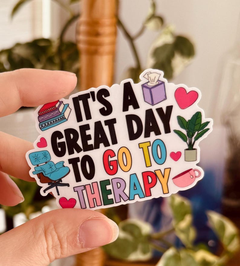 May include: A white sticker with the text "IT'S A GREAT DAY TO GO TO THERAPY" in various colours. The sticker features illustrations of books, a chair, a tissue box, a plant, a heart, and a coffee cup. The sticker is held by a hand.