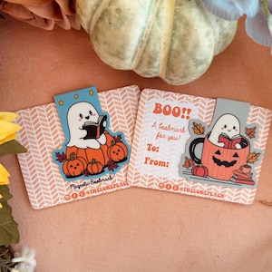 May include: Two Halloween-themed magnetic bookmarks. One features a ghost reading a book atop pumpkins, the other a ghost in a jack-o'-lantern mug. Both have the text "BOO!! A bookmark for you!" and "To: From:"