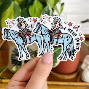 May include: Two blue horse stickers with a woman riding each horse. The woman has blonde hair and is wearing a blue and white outfit. The stickers have a white background with stars, moons, and the text "I NEED SPACE".