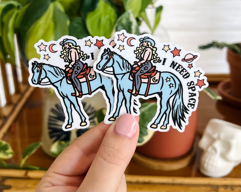 May include: Two blue horse stickers with a woman riding each horse. The woman has blonde hair and is wearing a blue and white outfit. The stickers have a white background with stars, moons, and the text "I NEED SPACE".