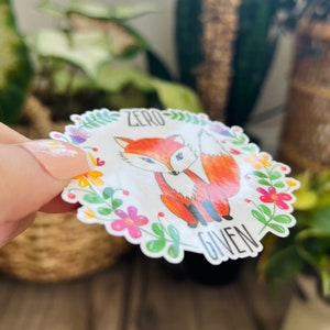 Pode incluir: Um adesivo branco com uma borda floral colorida. Uma raposa de desenho animado com um corpo laranja e uma cauda branca est&aacute; no centro do adesivo. O texto "ZERO GIVEN" est&aacute; escrito abaixo da raposa.