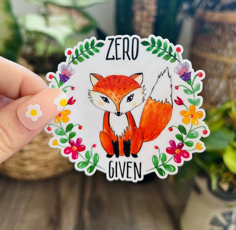 Pode incluir: Um adesivo branco com uma borda floral colorida. No centro, h&aacute; uma raposa de desenho animado com um corpo laranja, um peito branco e olhos pretos. O texto "ZERO GIVEN" est&aacute; escrito em preto abaixo da raposa.