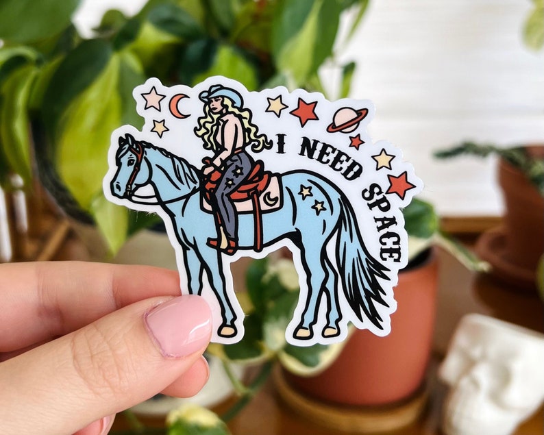 May include: A blue horse with a woman riding on it. The woman is wearing a cowboy hat and a blue shirt. The horse has a saddle and a bridle. The background is a starry sky with a crescent moon. The text "I NEED SPACE" is written on the sticker.