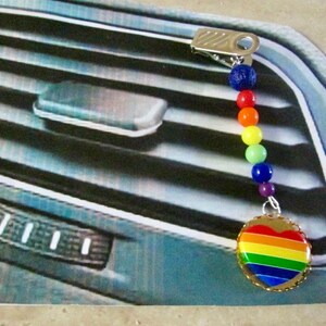 May include: A car air vent clip with a rainbow heart charm. The charm is a heart shape with a rainbow design and a gold-colored scalloped edge. The clip has a series of colorful beads in a rainbow pattern.