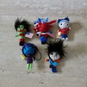 Puede incluir: Cinco muñecas de hilo de colores con diferentes peinados y accesorios. Las muñecas llevan ropa roja, azul, verde, morada y negra. Algunas de las muñecas sostienen banderas americanas.