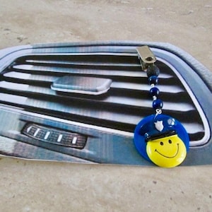 May include: A blue and yellow smiley face car air freshener with a police officer hat. The air freshener is hanging from a black beaded string attached to a silver clip.